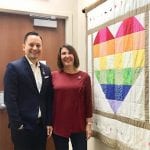 Two individuals smiling and standing next to a colorful quilt with a heart design.